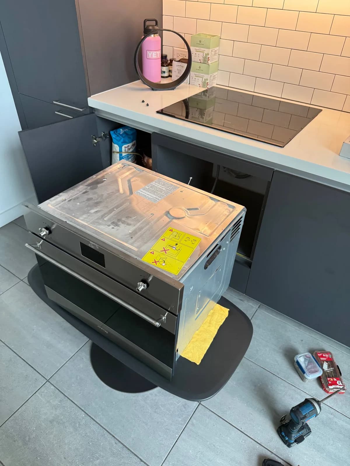 A stainless steel oven is pulled out from its cabinet space, resting on a mat, with tools on the floor nearby
