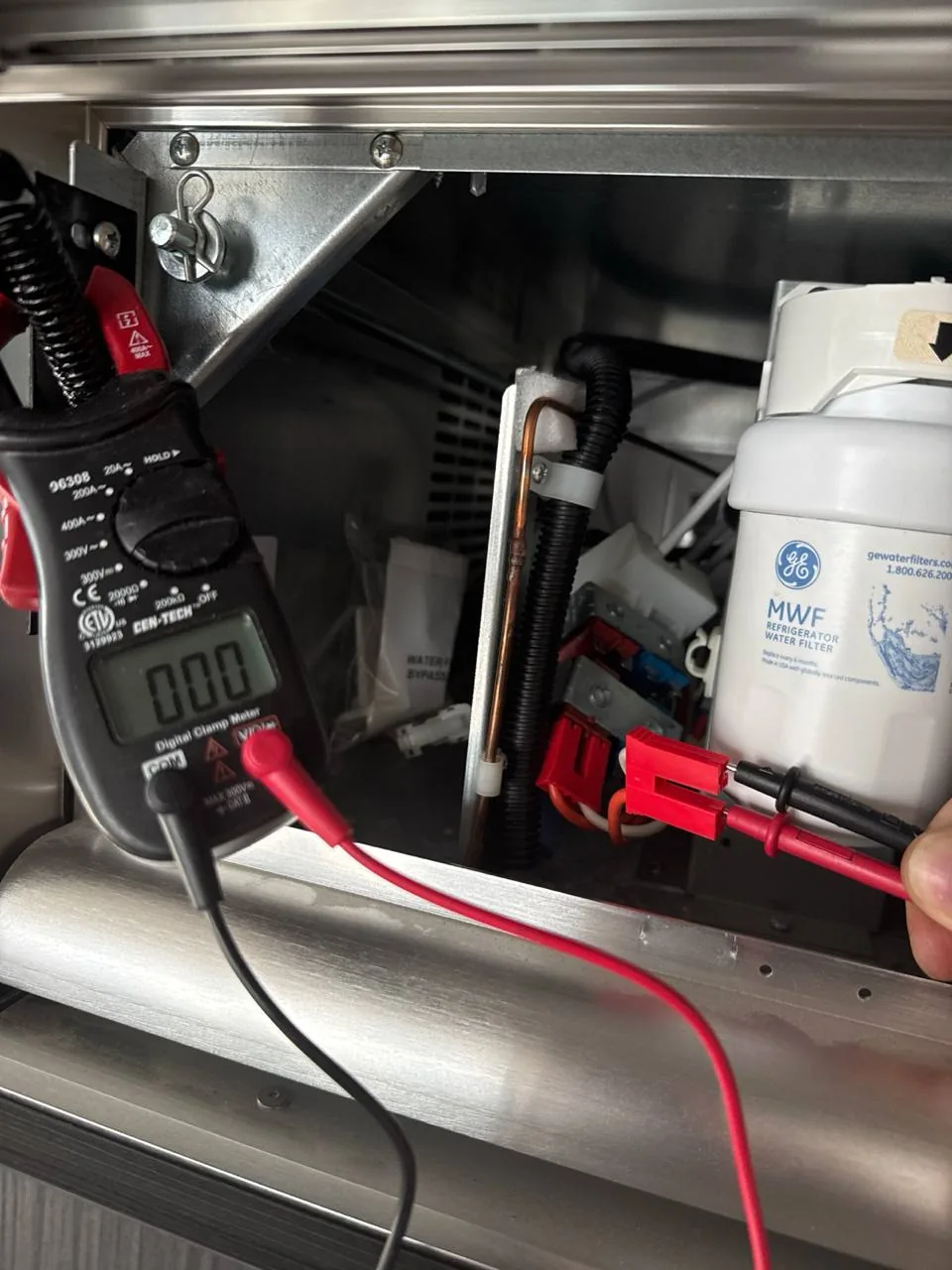 A technician is using a digital clamp meter to test components in a refrigerator, next to a GE water filter