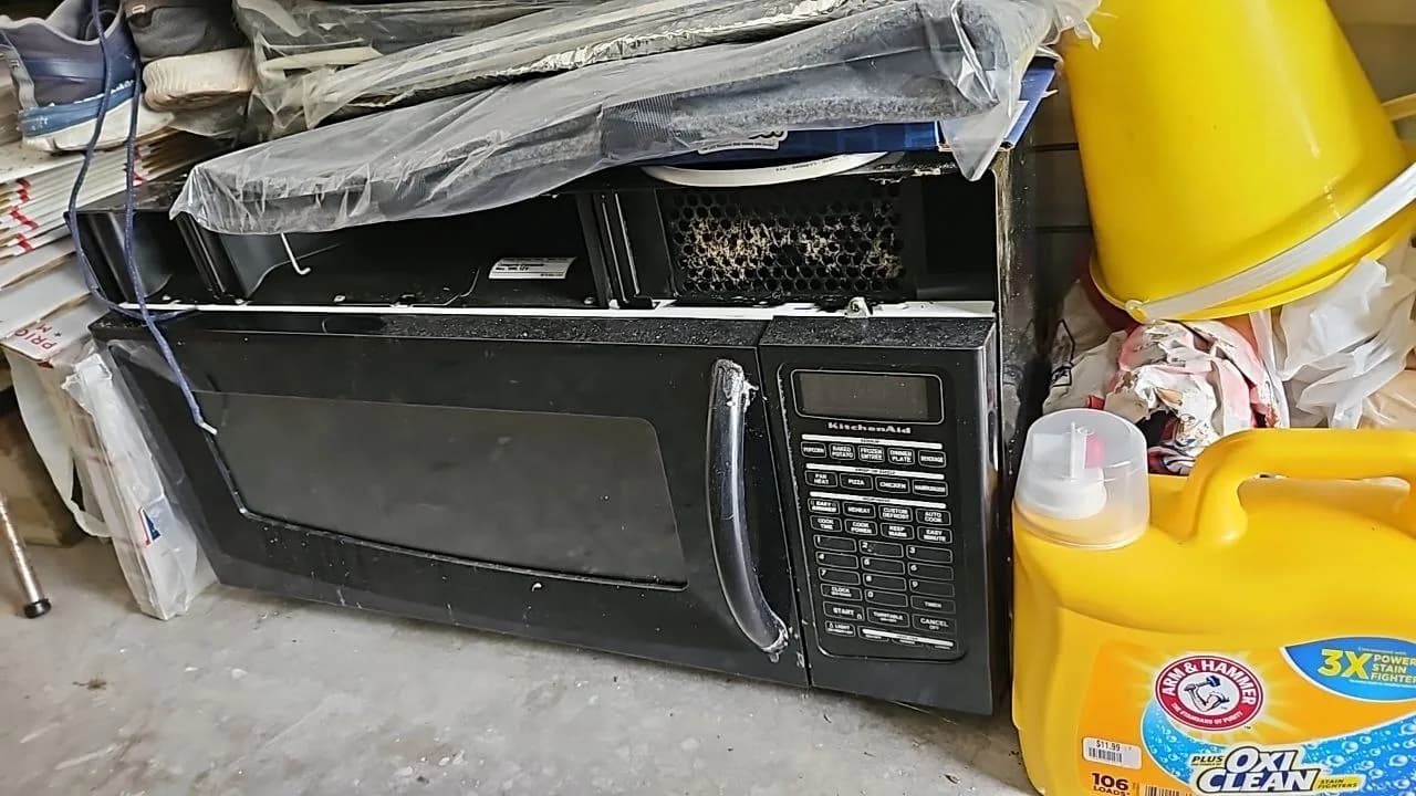 A black KitchenAid microwave oven sits on a floor, with part of its top cover removed, surrounded by laundry detergent and
