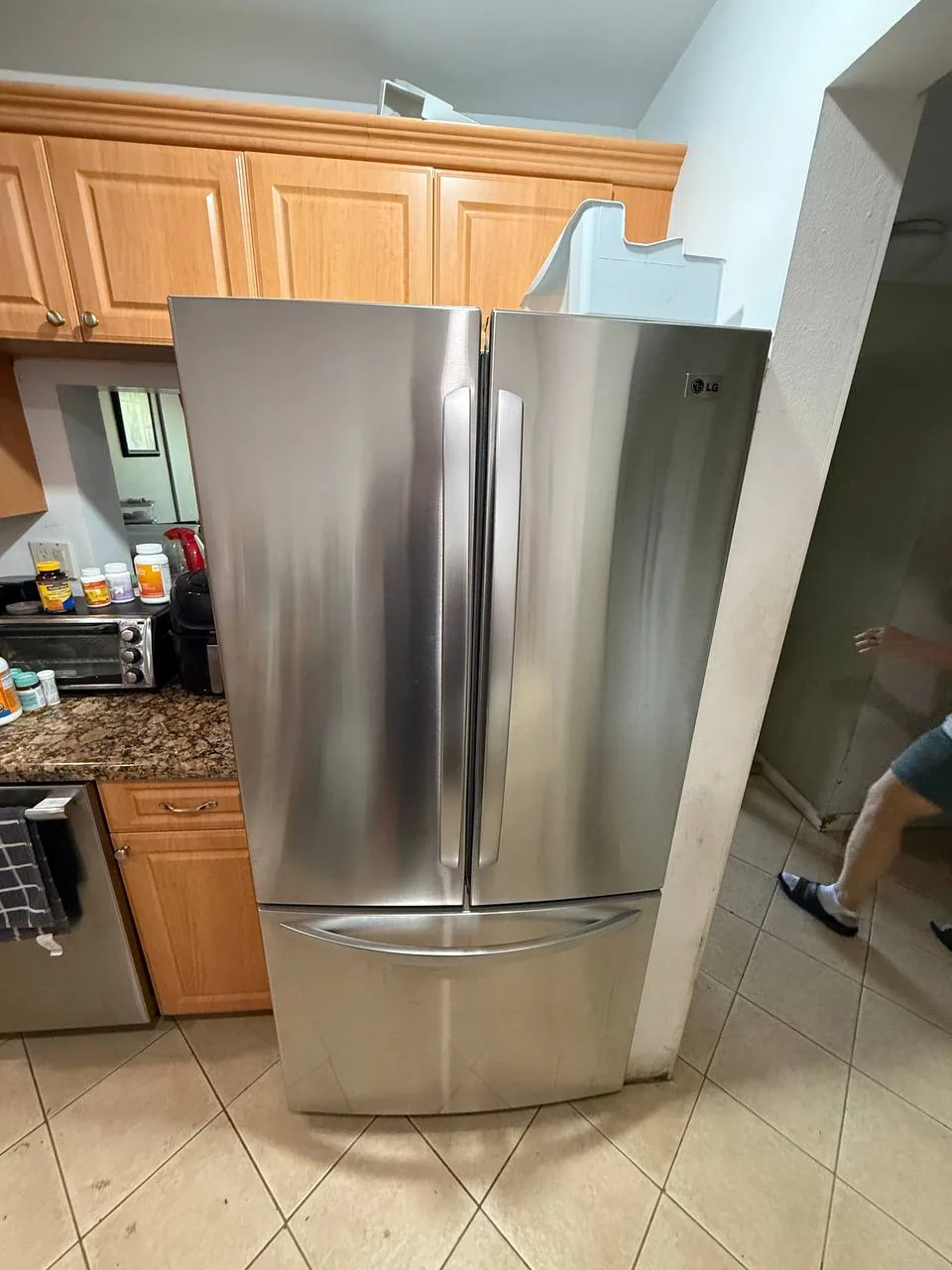 A stainless steel LG refrigerator in a kitchen setting with a technician's legs visible in the background
