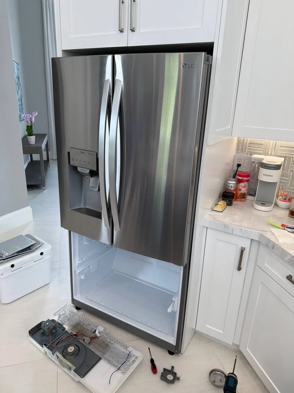 A stainless steel LG refrigerator with its freezer drawer open and internal components laid out on the floor during repair