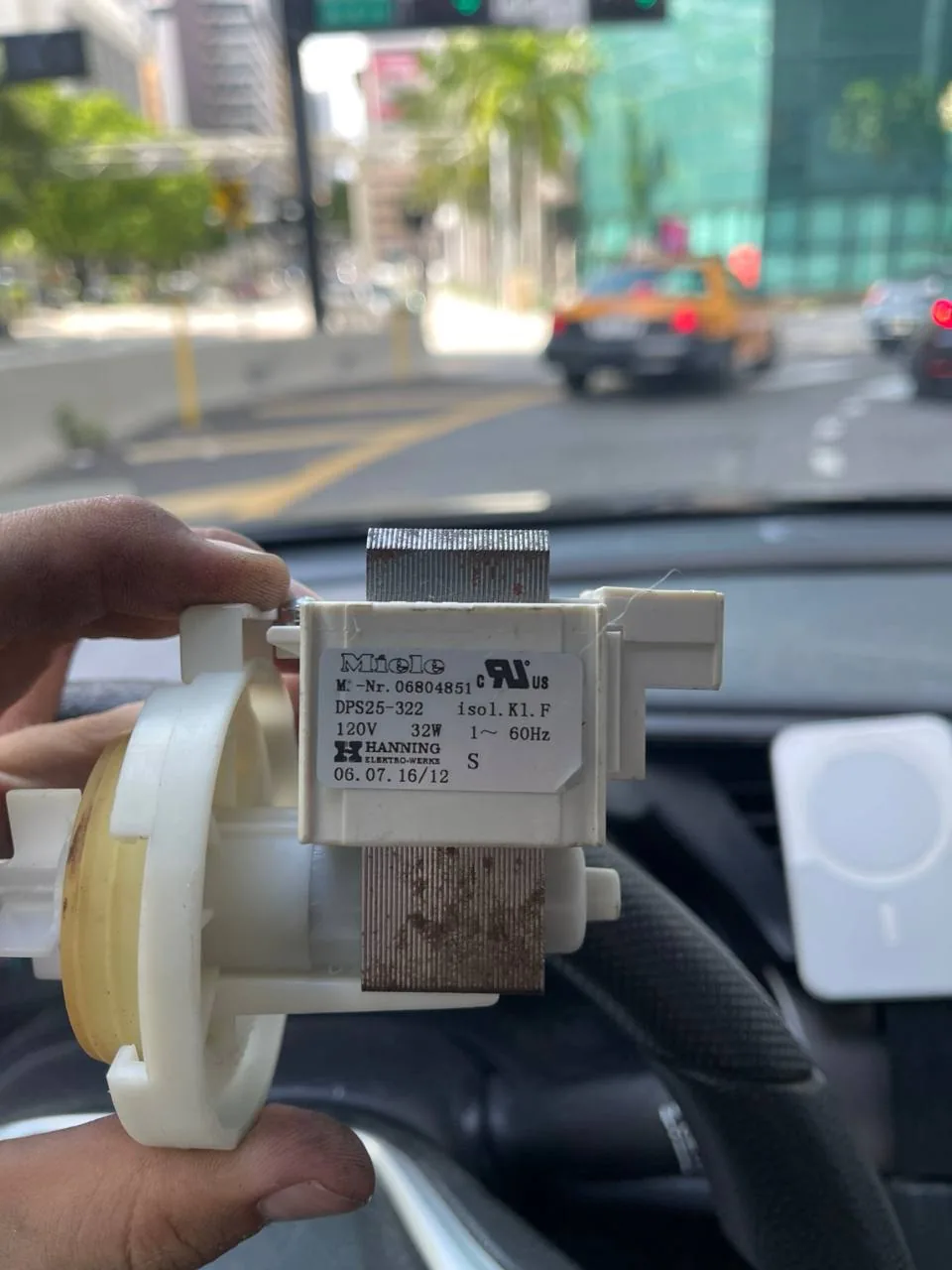 A technician holds a Miele part that appears to be a drain pump, with a street view blurred in the background