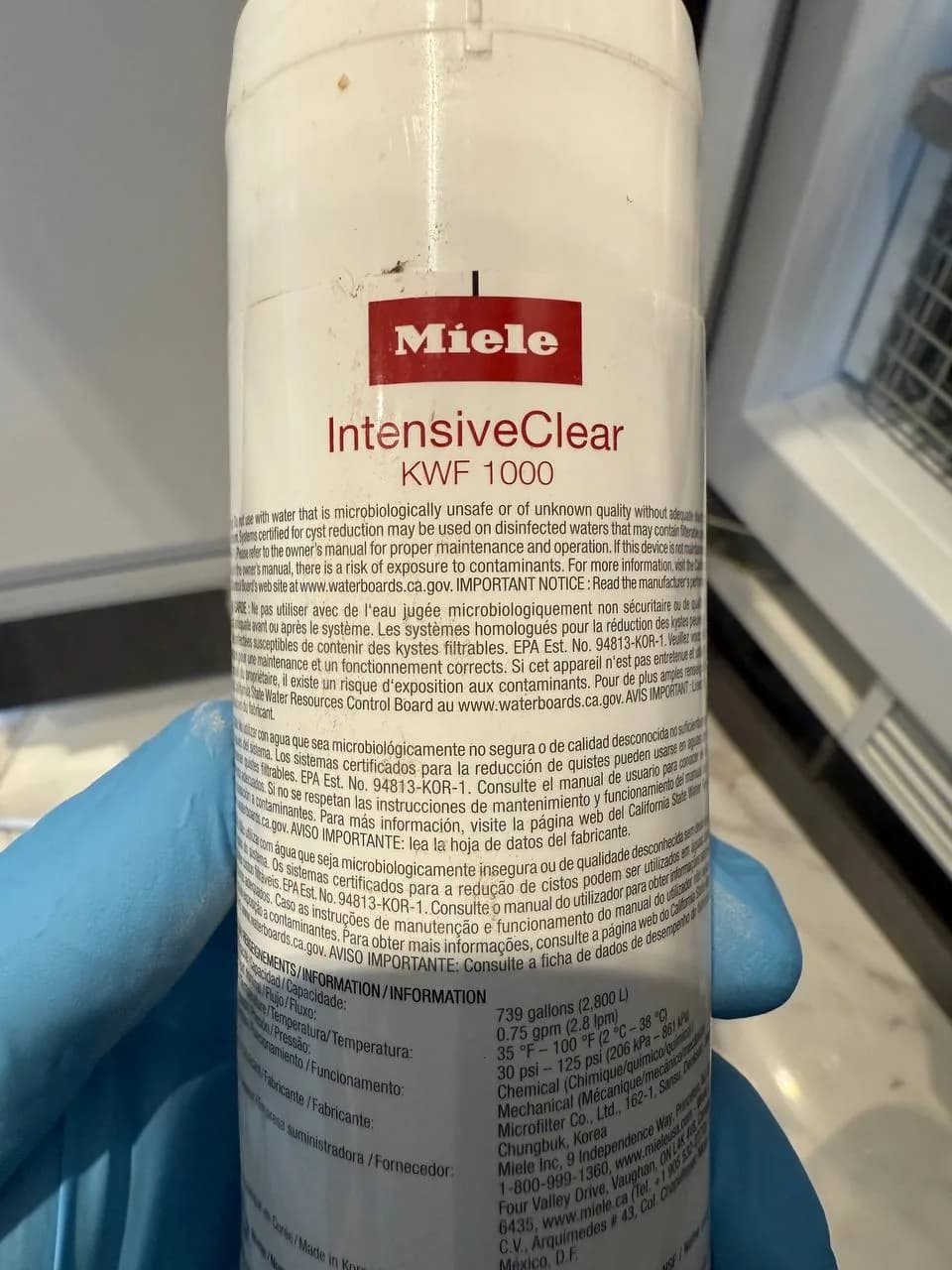 A Miele dishwasher water filter with text visible and a technician's gloved hand holding it