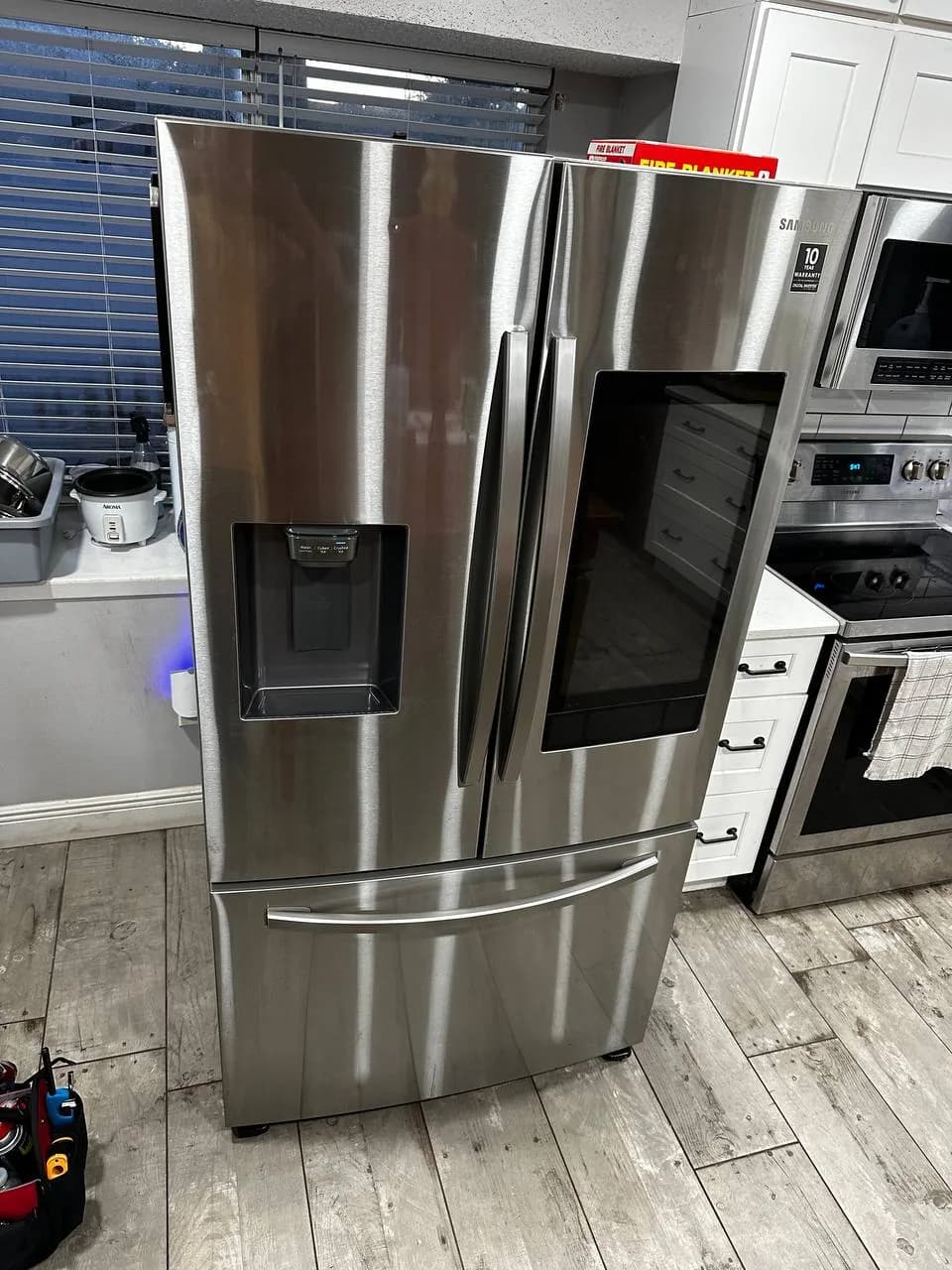A stainless steel Samsung refrigerator with a touch screen is prominently displayed in a kitchen setting, with a toolbox