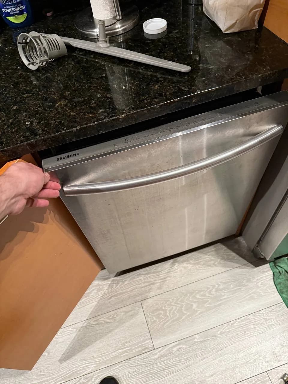 A technician is opening a Samsung dishwasher, with a spray arm and dish soap on the counter