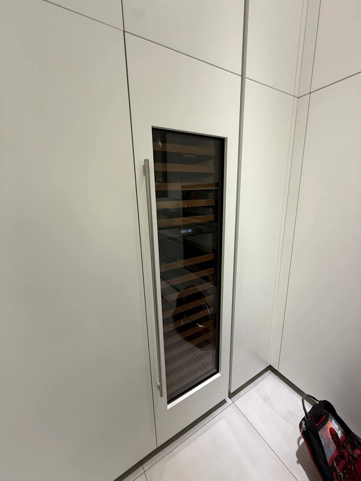 A modern Sub-Zero wine cooler is built into white cabinetry in a clean room, with a tool bag visible on the floor