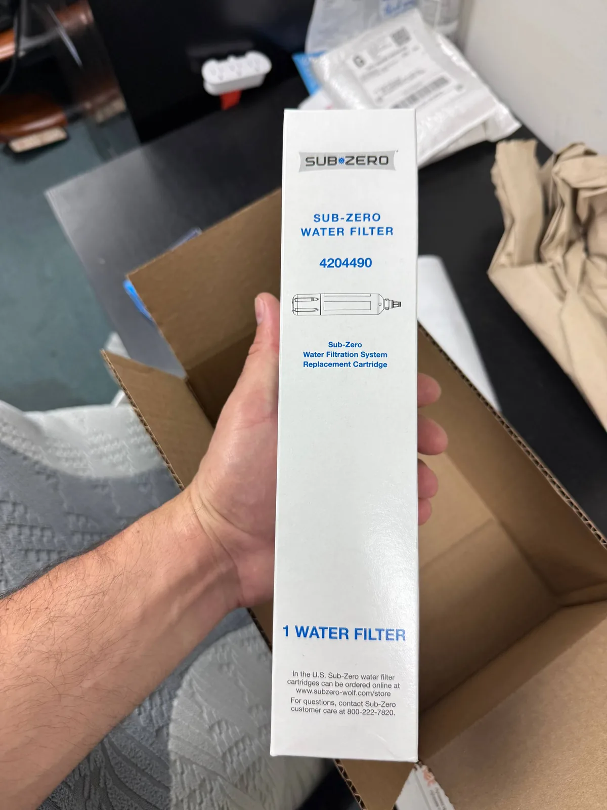 A technician holds a Sub-Zero water filter replacement cartridge box, with an open cardboard box visible in the background