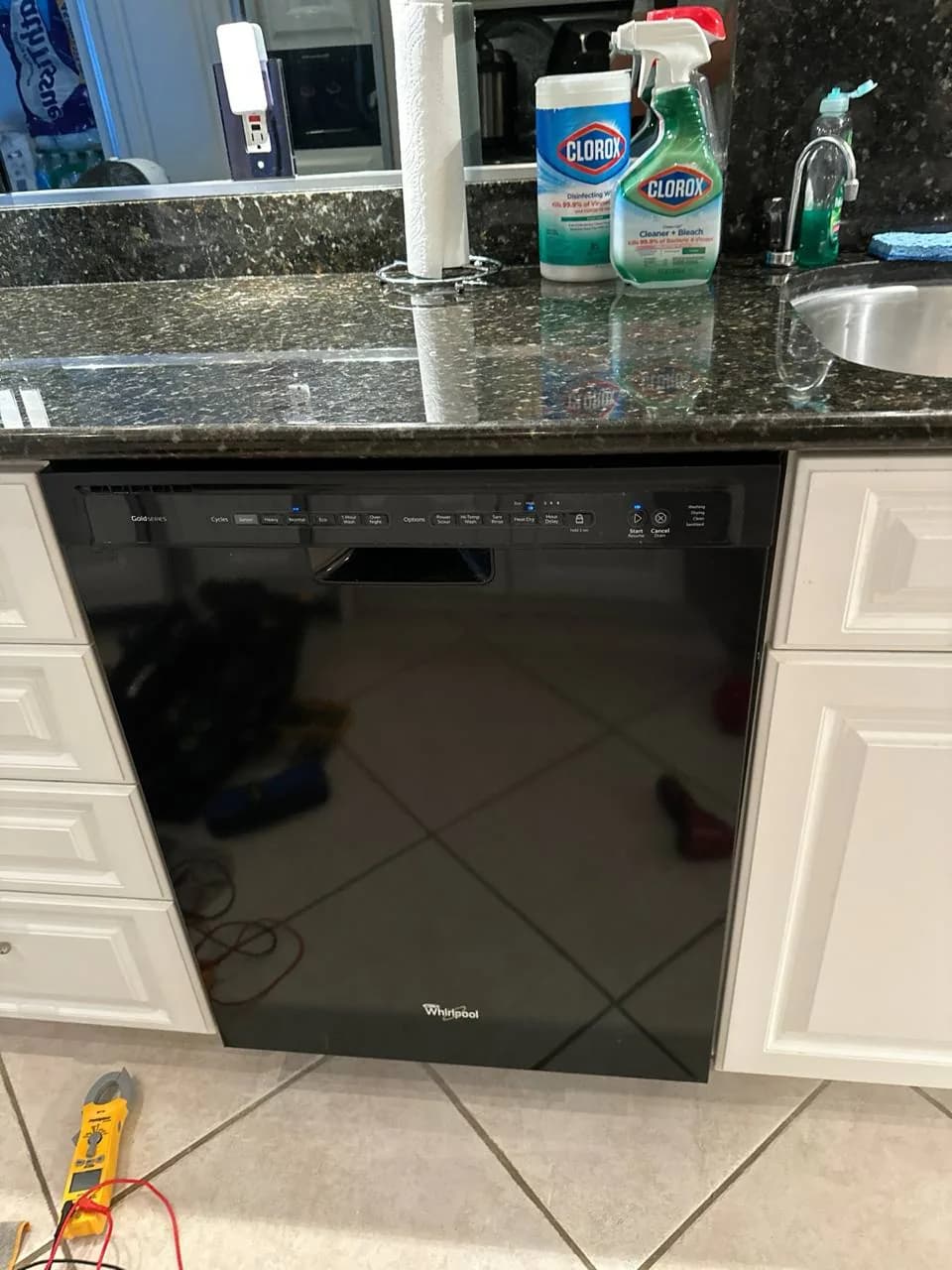A black Whirlpool dishwasher installed in a kitchen with a meter on the floor nearby