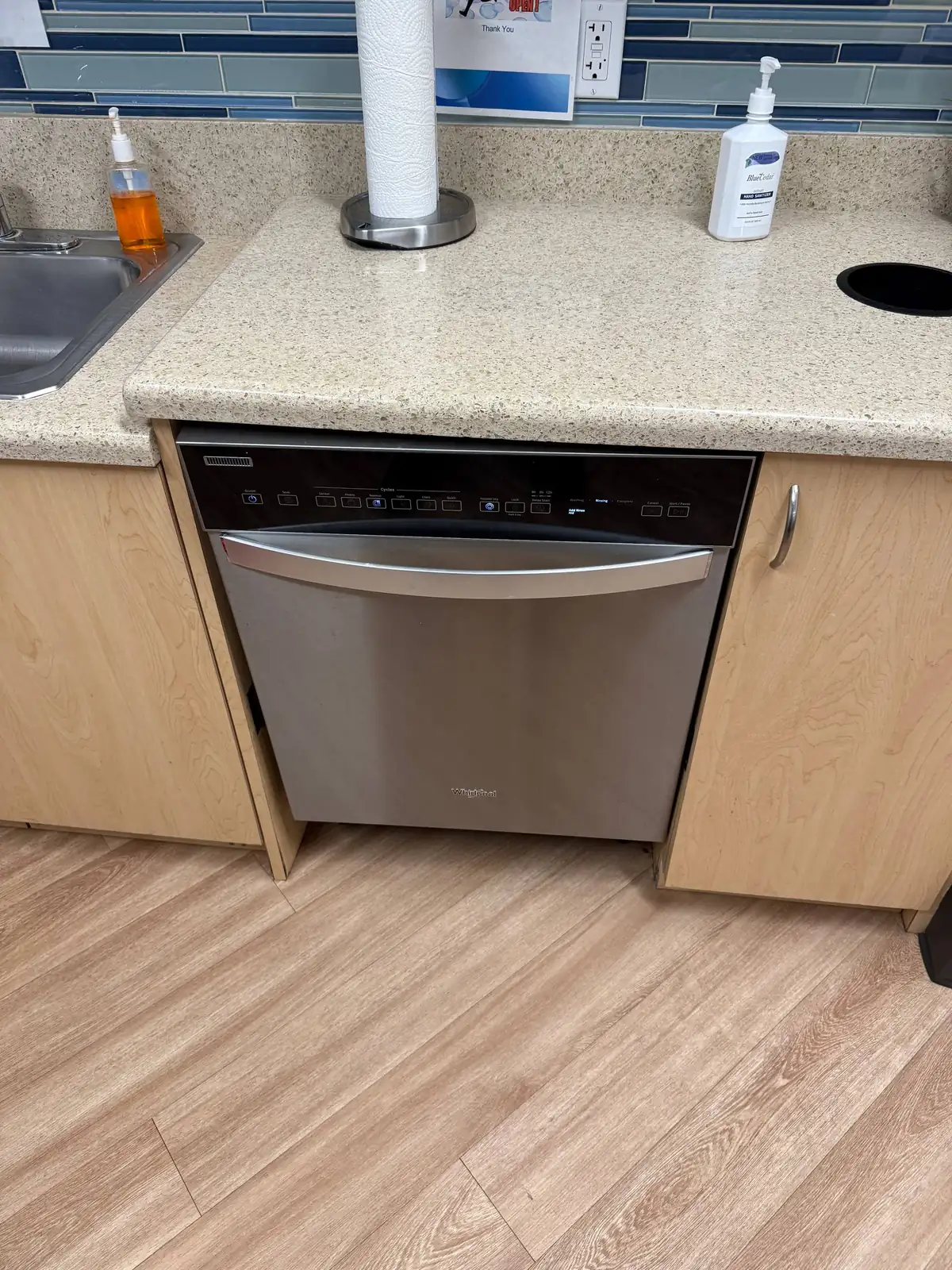A stainless steel Whirlpool dishwasher is installed under a counter with a sink and various bottles on top