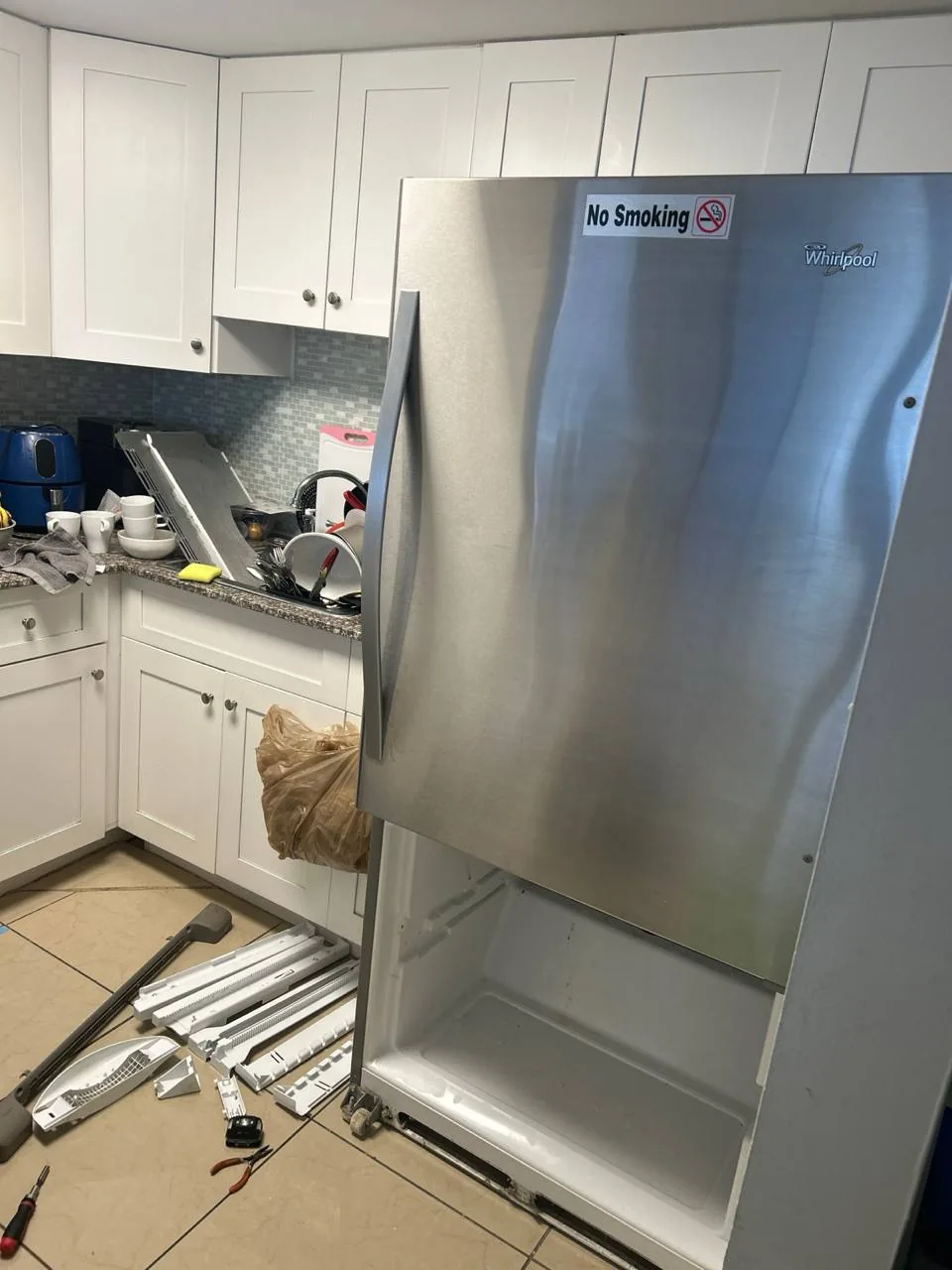 A stainless steel Whirlpool refrigerator with its bottom freezer door open, and refrigerator parts and tools are on the